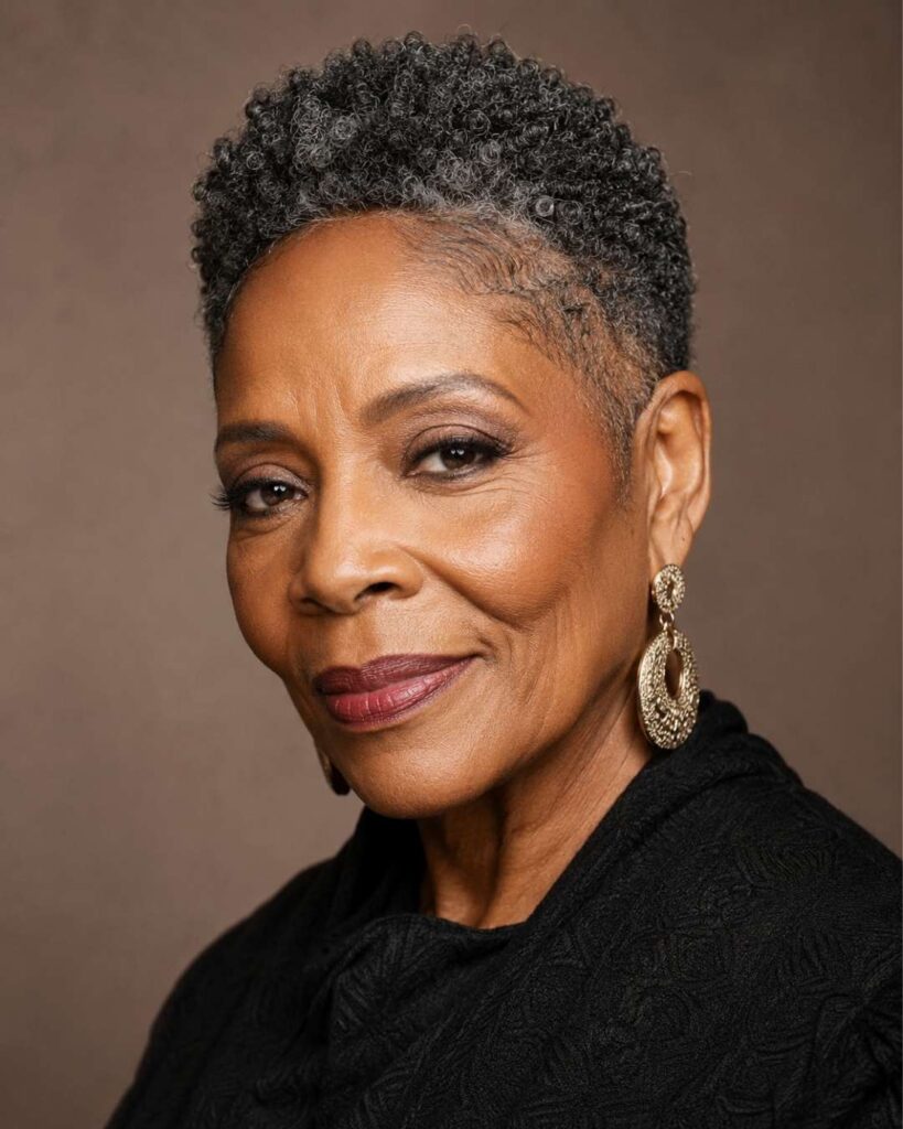 Elegant Black woman in her late 60s with a textured fade haircut and defined curls, wearing earrings in a warm studio.
